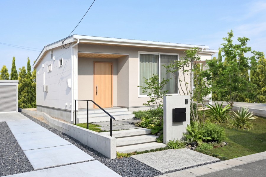 アイボリーとベージュの外壁、木製の玄関ドア、大きな窓、手入れの行き届いた前庭を備えたナチュラルな雰囲気の平屋。