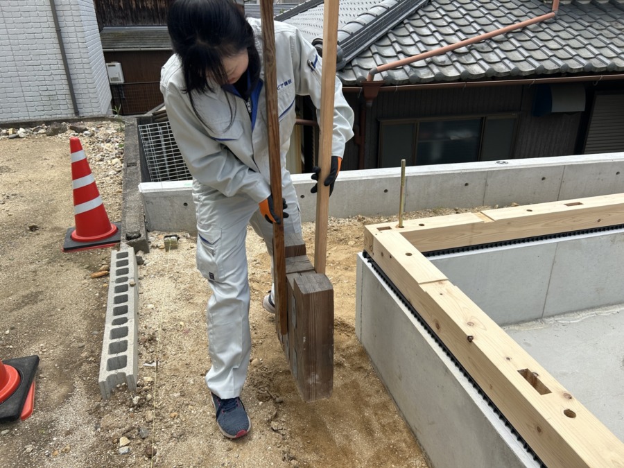 Construction worker in light gray coveralls and gloves lifts a wooden beam next to concrete formwork at a building site, with an orange traffic cone nearby.