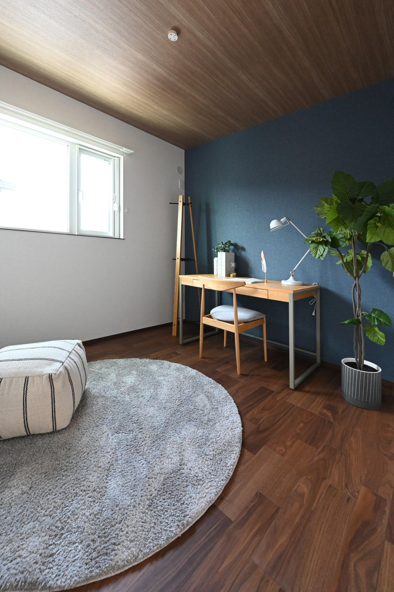 Modern home office corner with a wooden desk, chair, white desk lamp, blue accent wall, tall green plant, and a light gray round rug by a window.