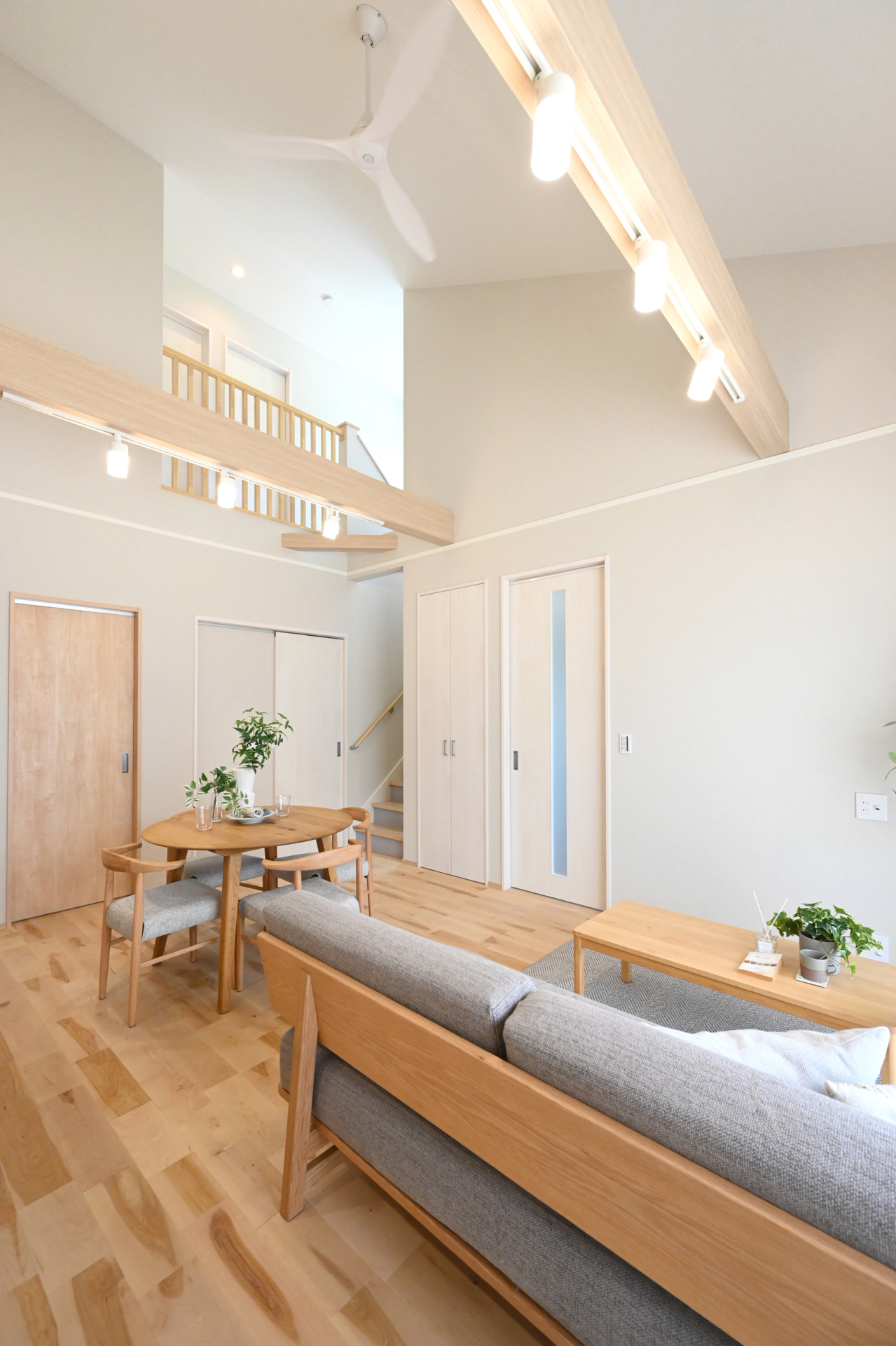 Bright open living area with light wood furniture, a round dining table, and a gray sofa under a high ceiling with warm lighting.
