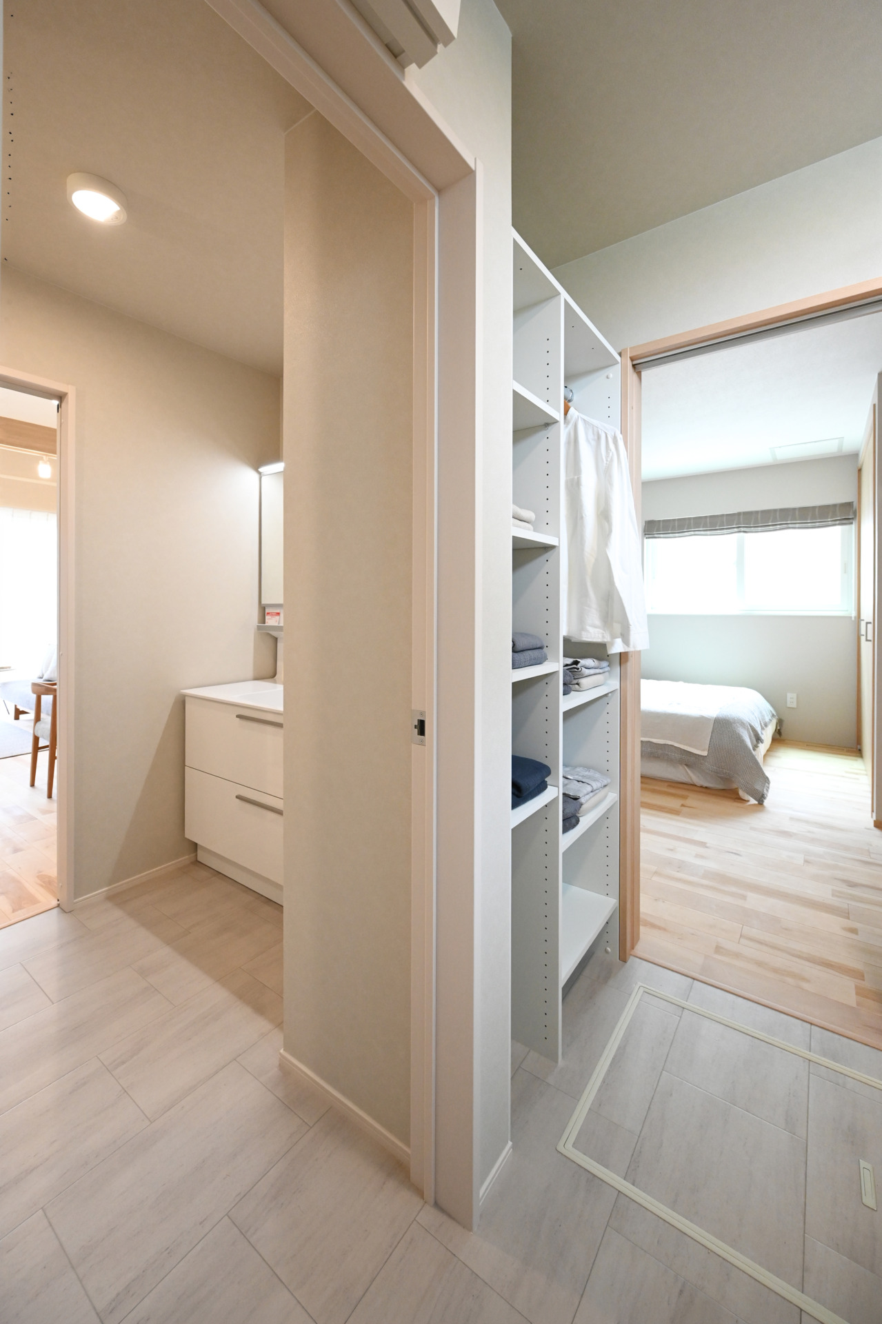 Closet with white shelves and folded towels, opening to a bright bedroom with a made bed and a bathroom vanity nearby.