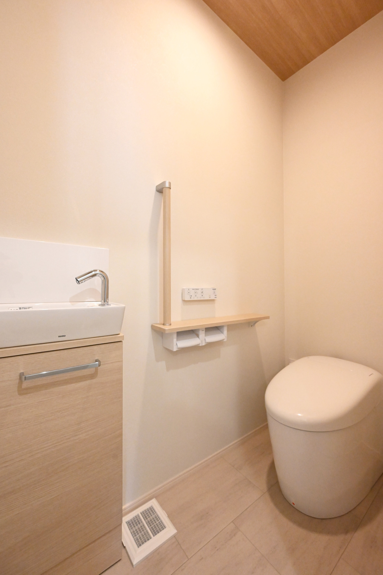 Compact, minimalist bathroom with a white toilet, light wood vanity and sink, and a vertical grab bar on the wall near the sink area.